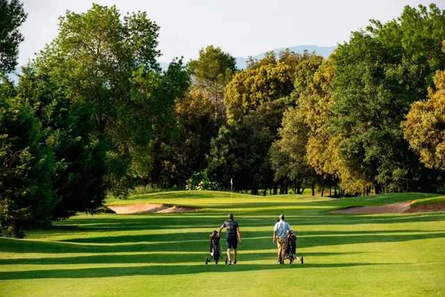 Dos golfistas caminando por el fairway de un campo de golf con árboles a ambos lados, bajo la luz del atardecer.