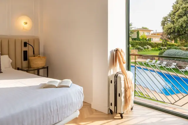 Habitación de hotel con cama grande vestida de blanco, mesita de noche con lámpara y cesta de mimbre, maleta blanca