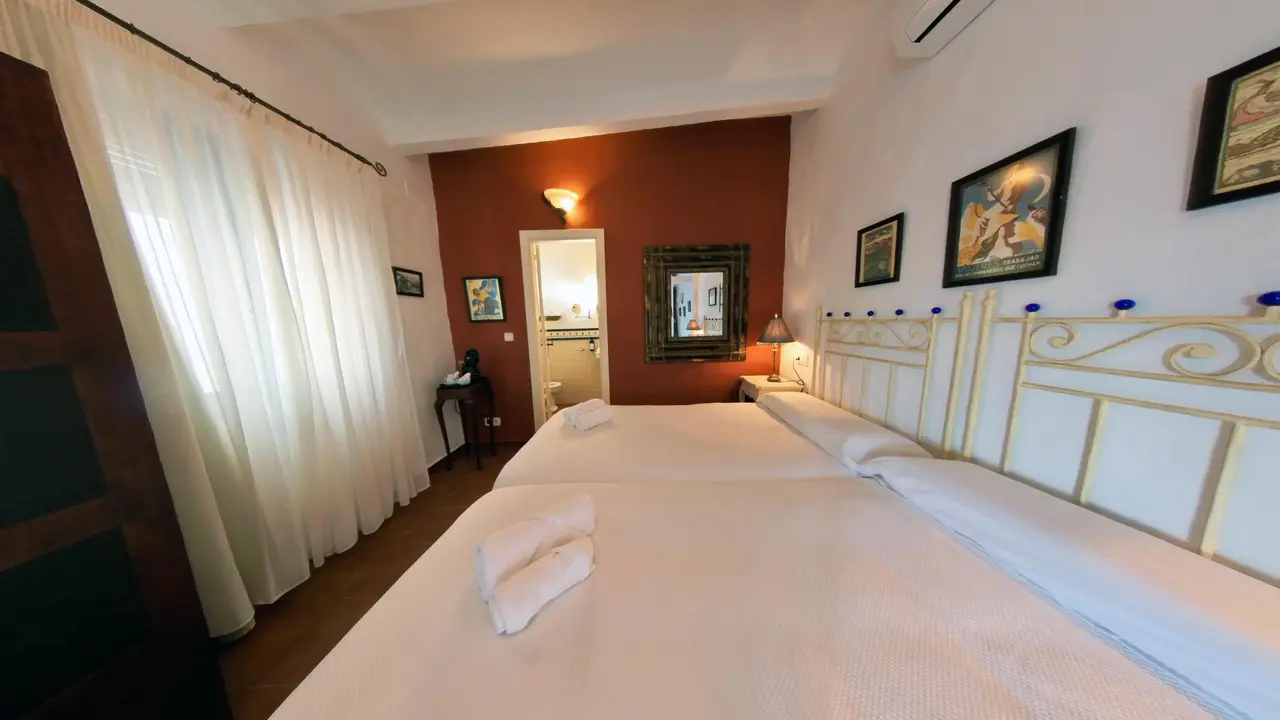 Habitación de hotel con dos camas individuales separadas, cabeceros blancos con detalles dorados, colchas blancas