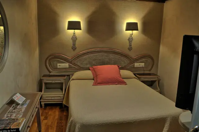 Vista de una suite con cama doble, cabecero de madera tallada, dos mesitas de noche con lámparas de pared, un cojín