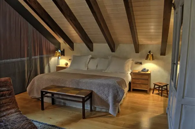 Vista de una suite junior con cama grande vestida con edredón y múltiples almohadas, cabecero de madera, mesitas