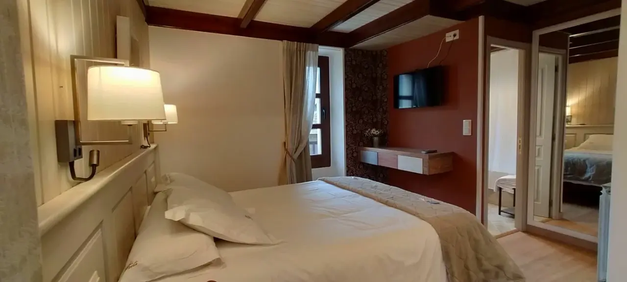 Vista de una suite junior con una cama grande vestida de blanco, cabecero de madera clara, lámparas de pared, vigas