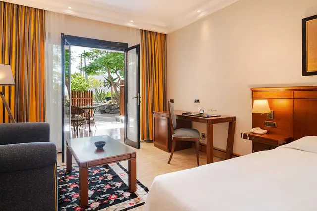 Vista de una suite de hotel con cama grande a la derecha, escritorio de madera con silla, sofá gris y mesa de centro