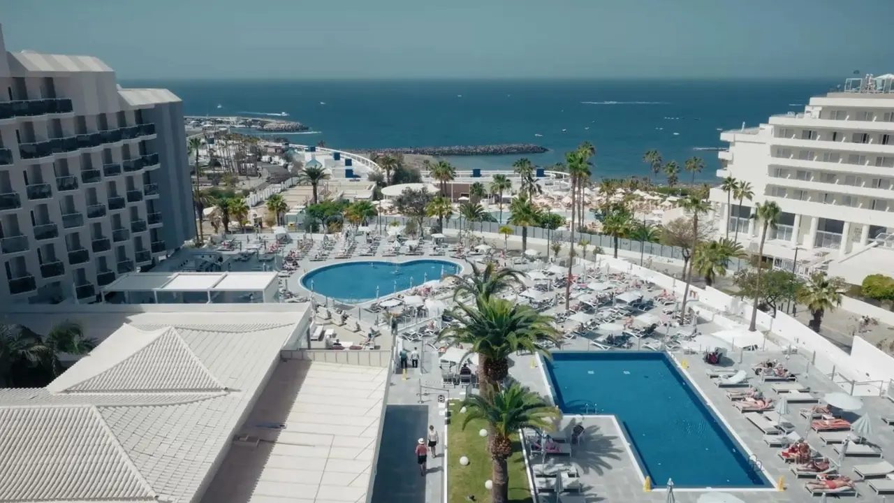 Vista panorámica de la zona de piscinas y solárium de un resort costero, con dos piscinas rectangulares y una en forma