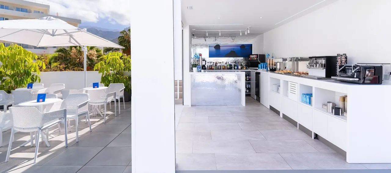 Terraza exterior con mesas y sillas blancas bajo sombrillas, junto a un bar interior moderno con encimera blanca