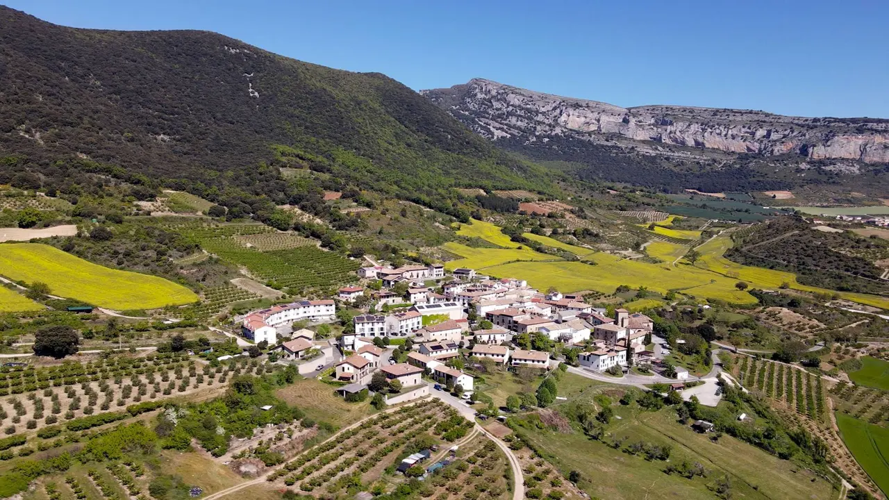 Vista aérea de un pueblo rural rodeado de campos de cultivo y montañas al fondo bajo un cielo despejado.