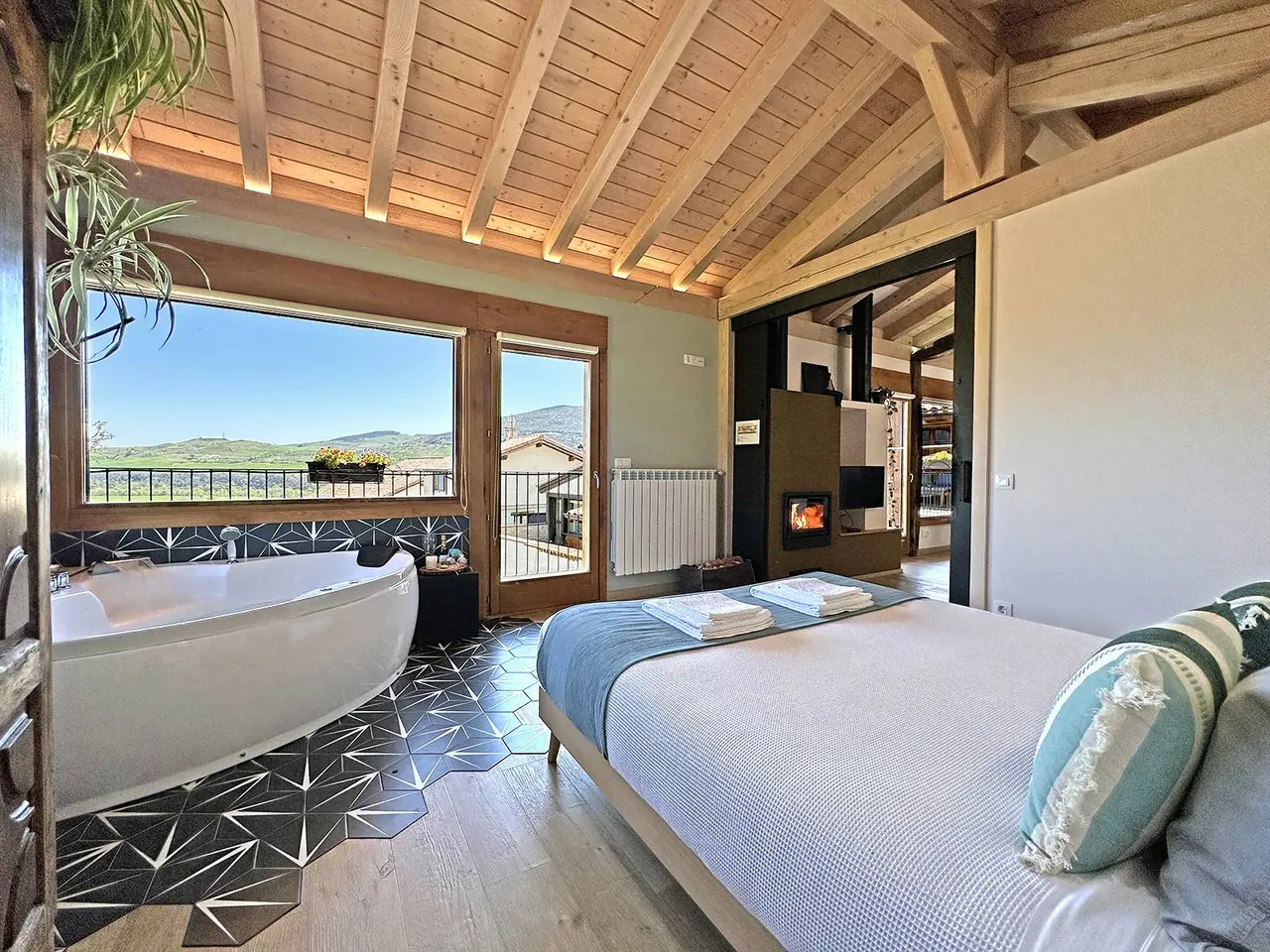 Habitación con jacuzzi blanco junto a una ventana grande con vistas a las montañas, cama grande con colcha blanca