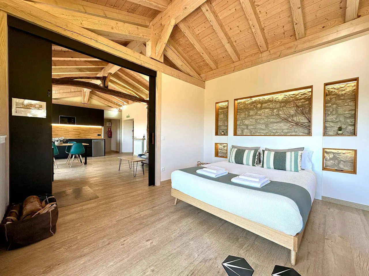 Vista de una habitación amplia con cama doble, suelo de madera clara y techo abuhardillado con vigas de madera.