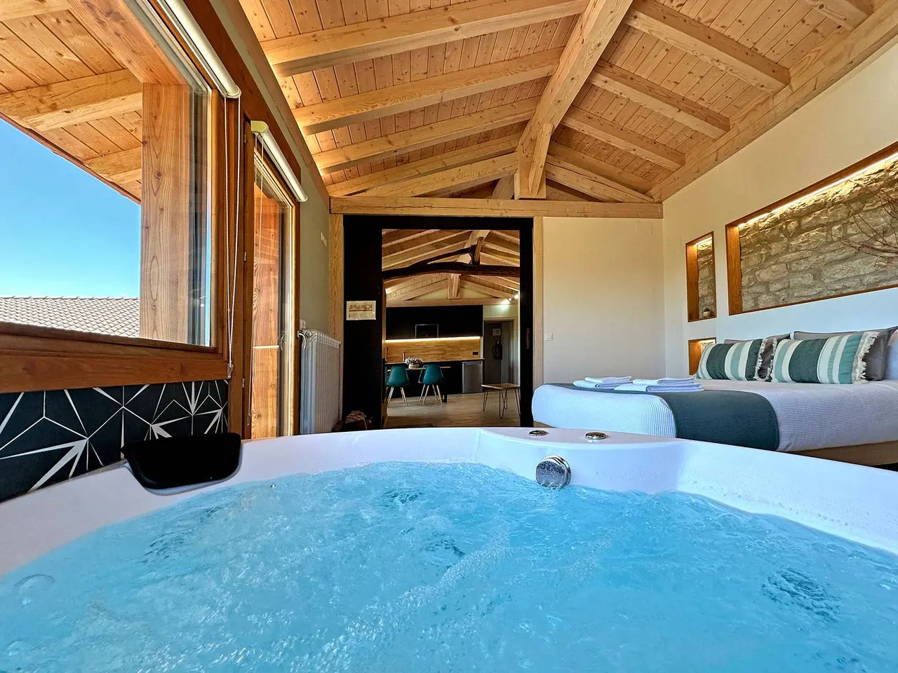 Jacuzzi blanco con agua burbujeante en primer plano dentro de una habitación con techo de madera, cama doble al fondo