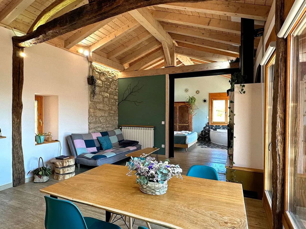 Vista de un salón con mesa de comedor de madera, sofá multicolor y vigas de madera en el techo.