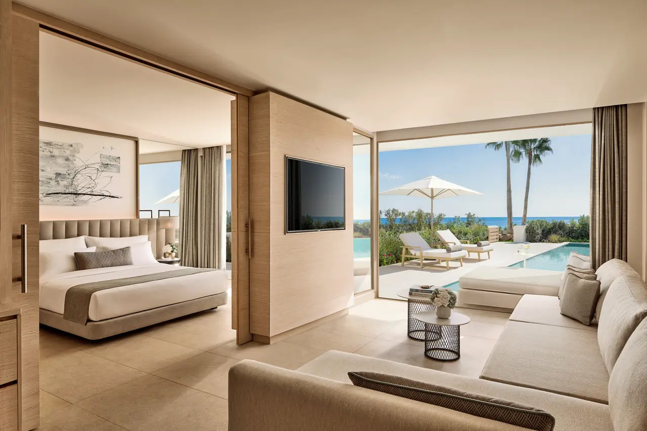 Vista interior de una suite con salón, sofá, mesa de centro, televisor en la pared y vistas directas a una piscina