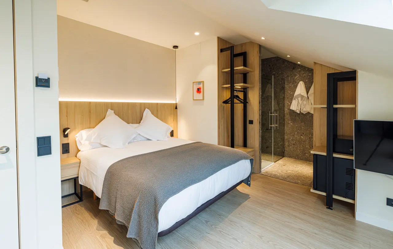 Vista de una habitación suite con cama doble grande vestida con sábanas blancas y manta gris, cabecero de madera