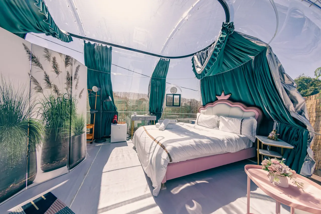 Habitación interior de burbuja transparente con cama doble de color rosa, dosel verde y vistas al exterior soleado.