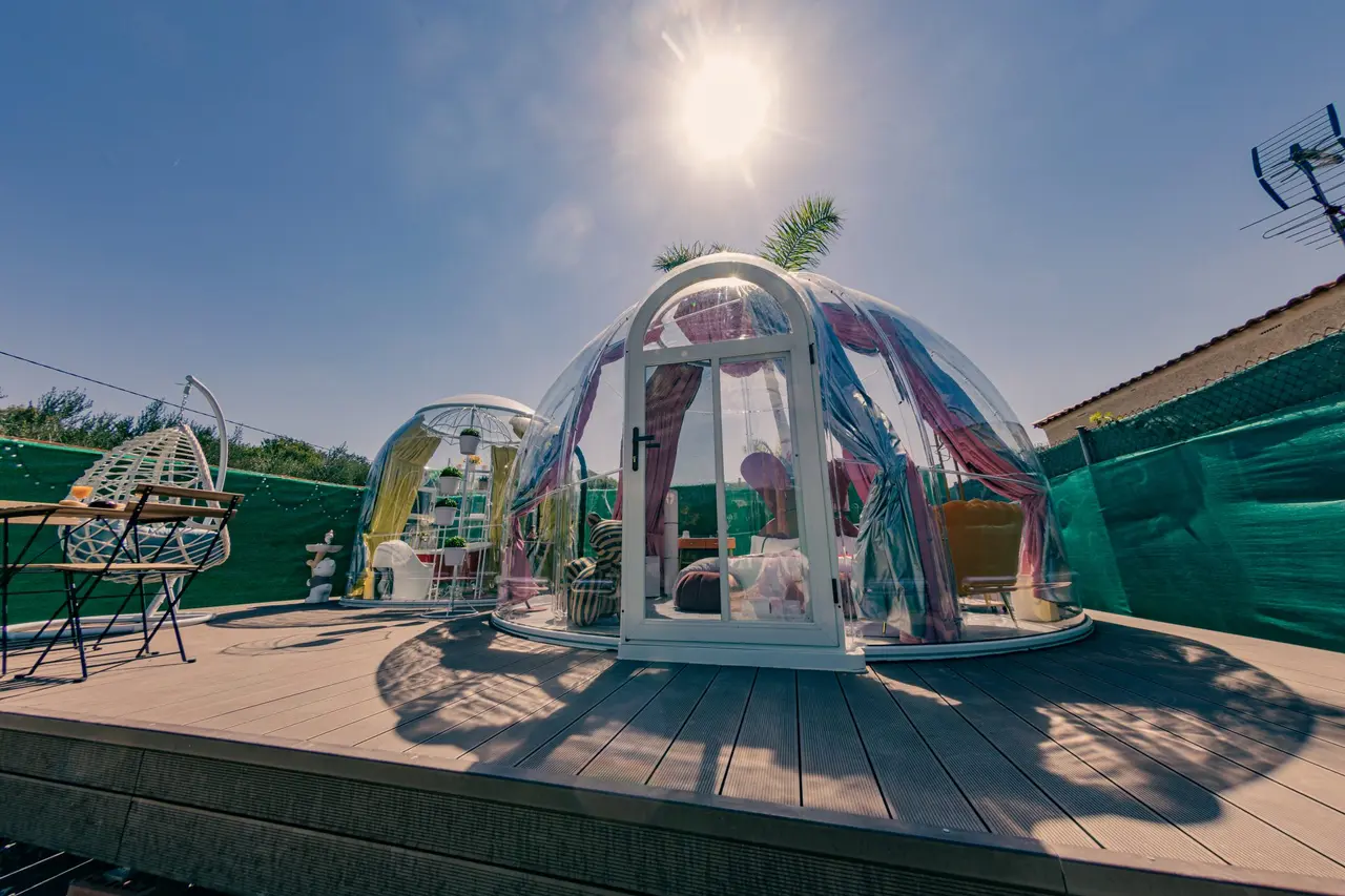 Burbuja transparente de glamping con cortinas de colores en una terraza de madera bajo el sol.