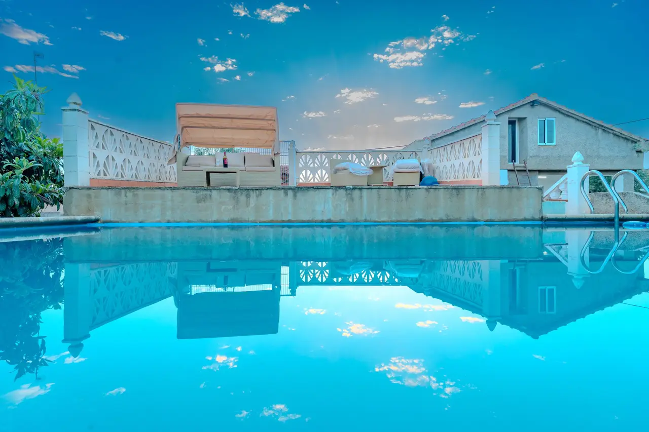 Piscina rectangular con agua azul reflejando el cielo, rodeada de una terraza con barandilla blanca y mobiliario