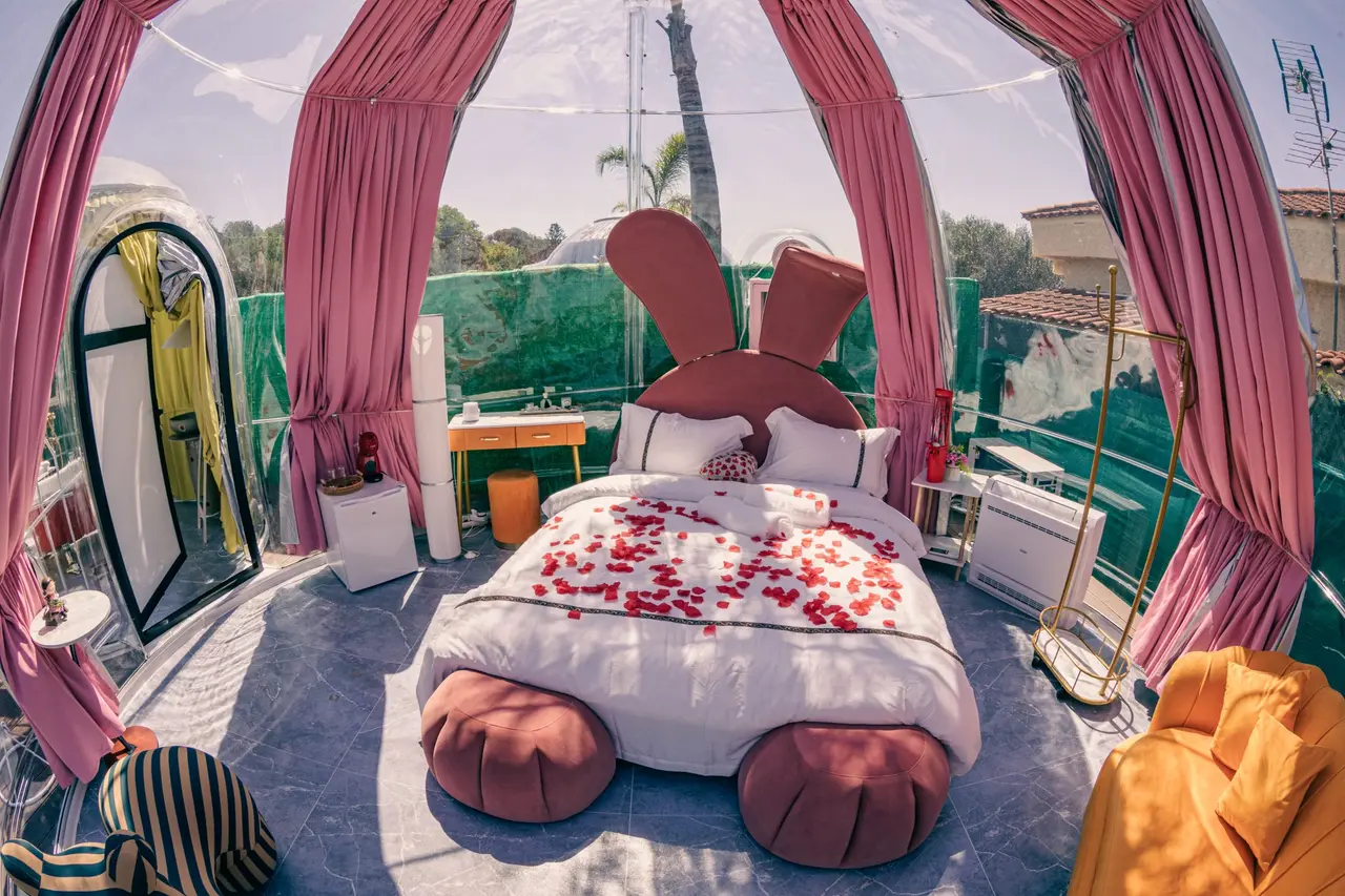 Habitación en burbuja transparente con cama redonda en forma de conejo, pétalos rojos y cortinas rosas, con vistas