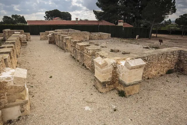 Ruinas arqueológicas de muros de piedra en un yacimiento al aire libre con grava en el suelo y edificios al fondo.