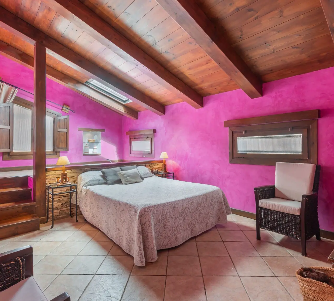 Habitación con paredes magenta, techo de vigas de madera, cama grande con colcha clara, sillón de mimbre, suelo