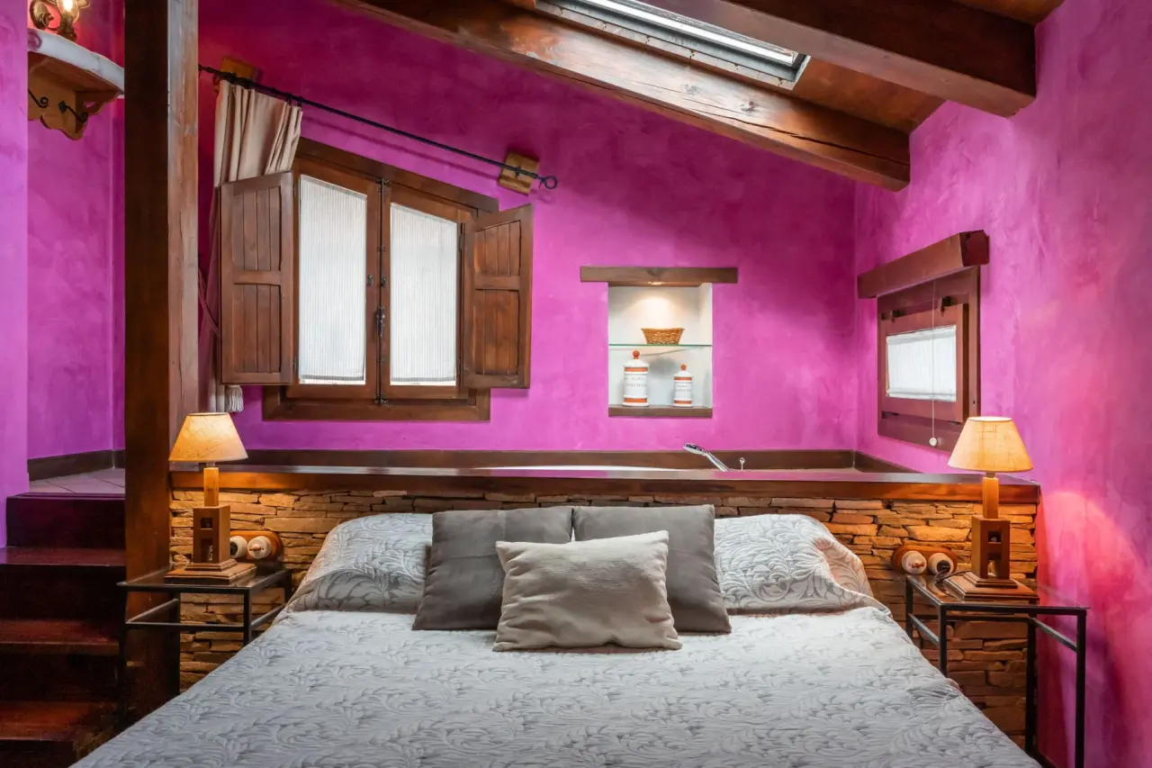 Habitación con paredes magenta, techo con vigas de madera y claraboya. Una gran cama con cabecero de piedra y cojines