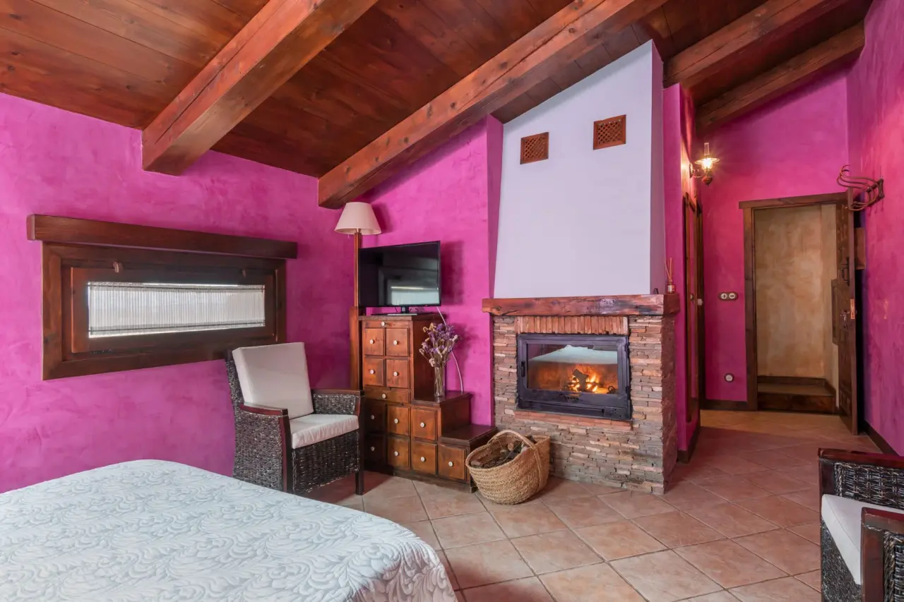Interior de una habitación con paredes magenta, techo de vigas de madera, cama con colcha blanca en primer plano