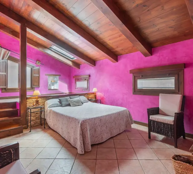 Habitación con paredes magenta, techo de vigas de madera, cama doble con colcha clara, sillón de mimbre, ventanas