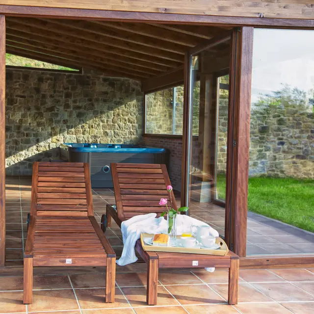 Jacuzzi exterior cubierto con agua azul, situado bajo un porche de madera junto a una pared de piedra.