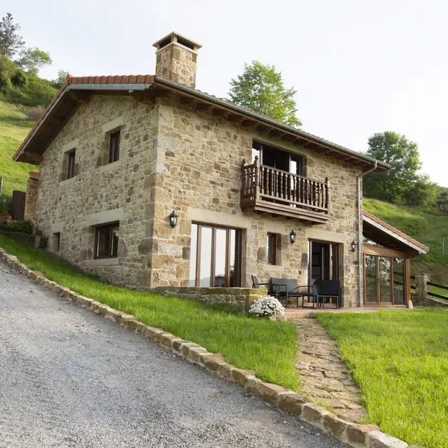 Vista exterior de una casa rural de piedra con dos plantas, tejado a dos aguas, chimenea, balcón de madera y terraza