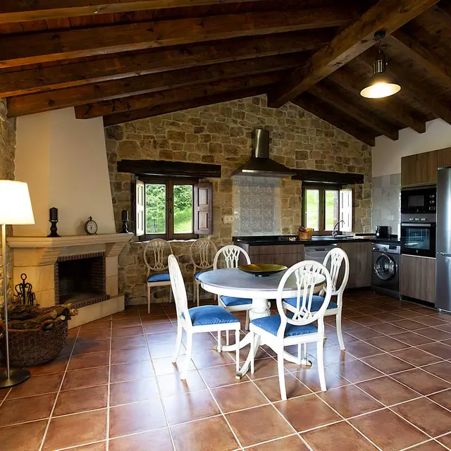 Interior de una casa rural con suelo de terracota, mesa de comedor redonda con sillas blancas y cojines azules, cocina