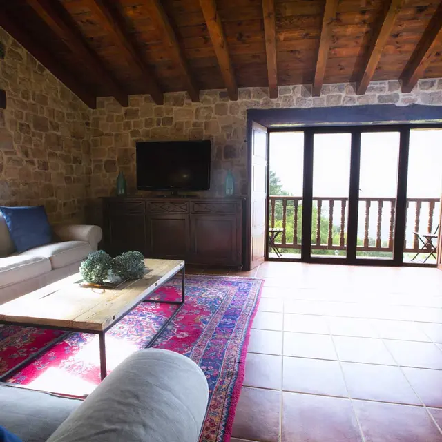 Interior de una sala de estar rústica con paredes de piedra, techo de vigas de madera, sofá claro, mesa de centro