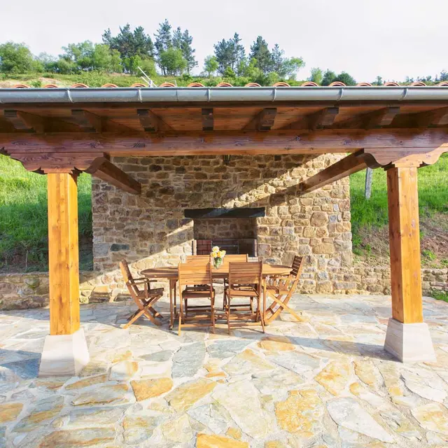 Porche exterior cubierto con suelo de piedra irregular, mesa y sillas de madera, frente a una pared de piedra