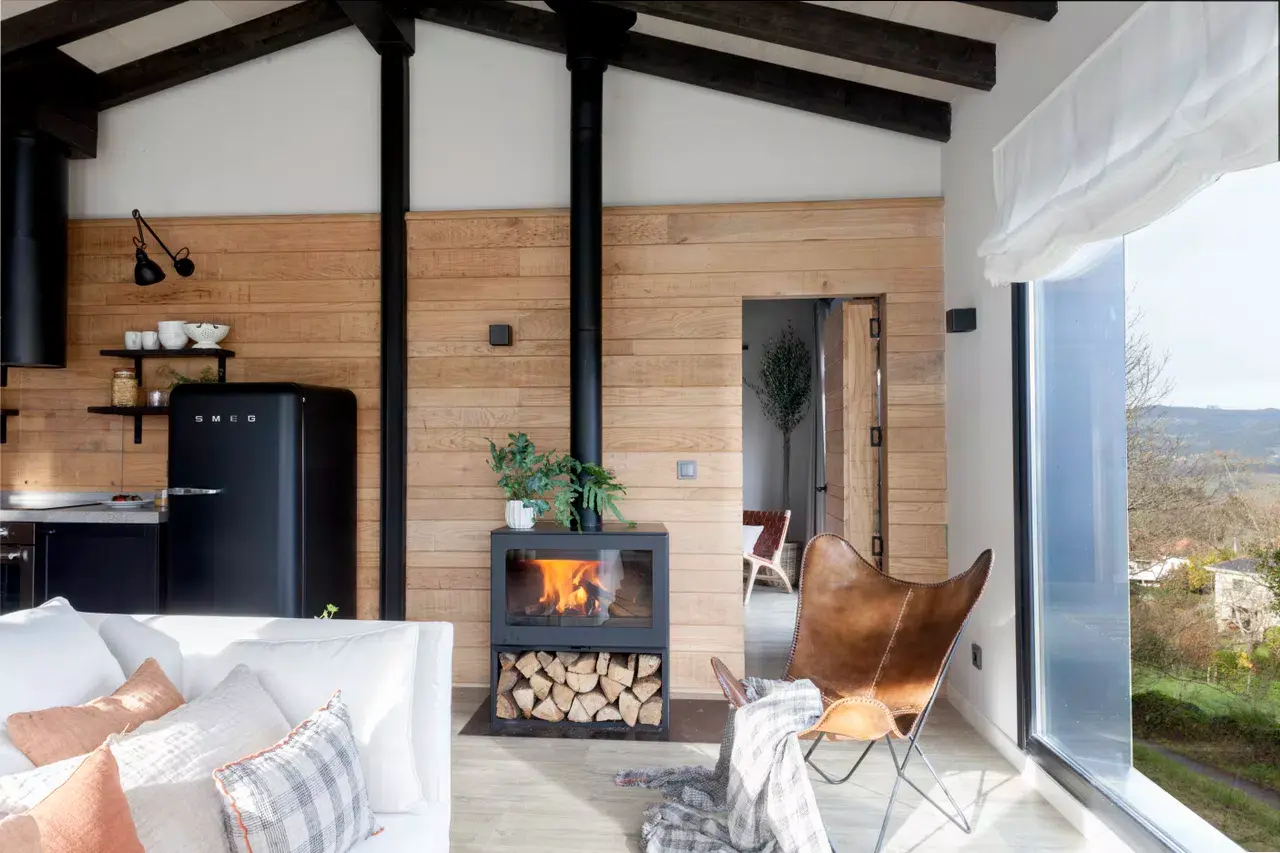 Interior de una cabaña con paredes de madera clara, vigas oscuras en el techo, sofá blanco con cojines, chimenea