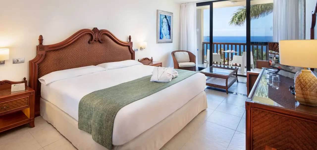 Habitación de hotel con cama king size de madera tallada, colcha blanca y verde, mesitas de noche, escritorio