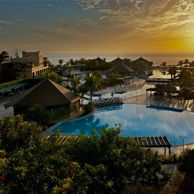 Vista panorámica de una gran piscina con agua azul en un complejo turístico al atardecer, rodeada de vegetación