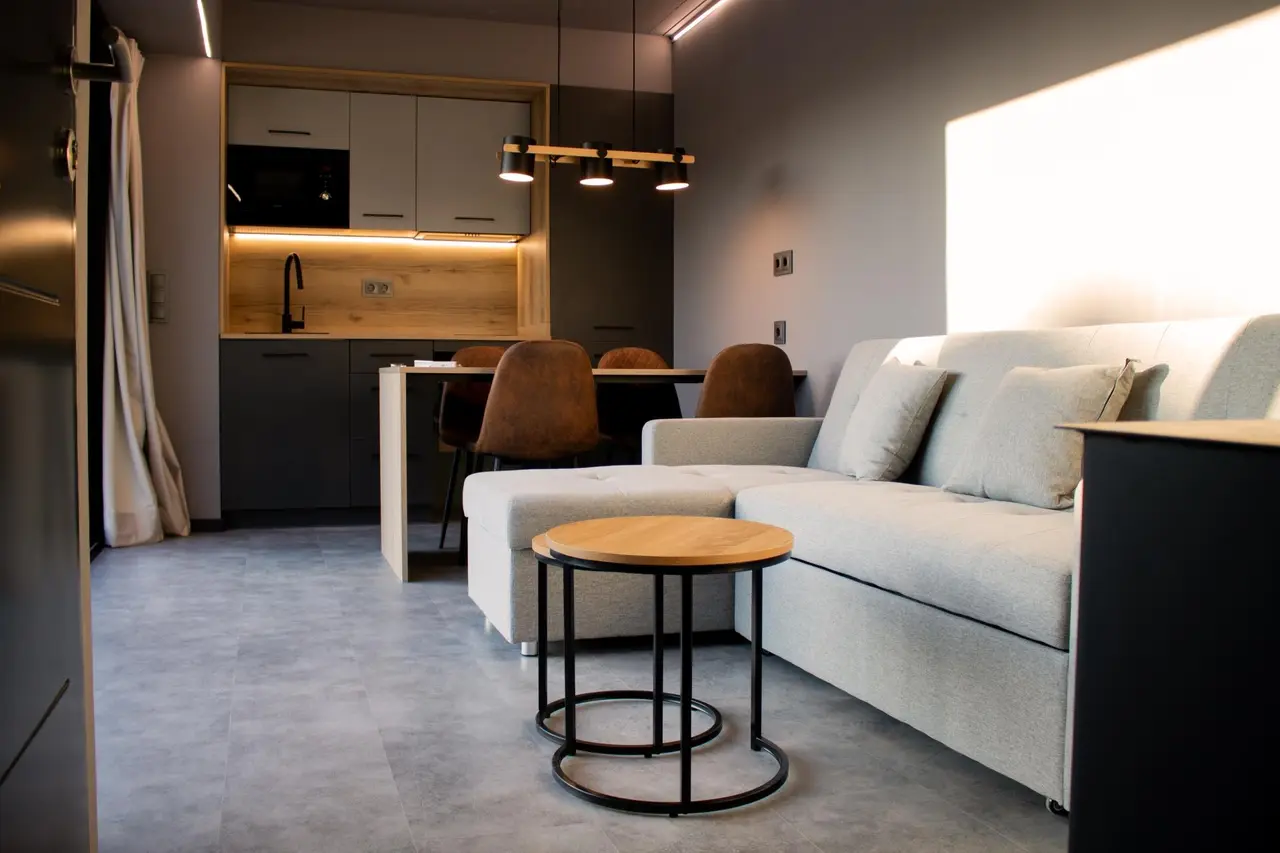 Vista de una sala de estar con sofá claro en forma de L, mesa auxiliar redonda de madera y metal, y cocina americana