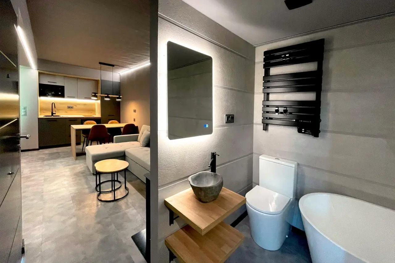 Baño moderno con bañera blanca de hidromasaje, inodoro, lavabo de piedra sobre encimera de madera, espejo iluminado