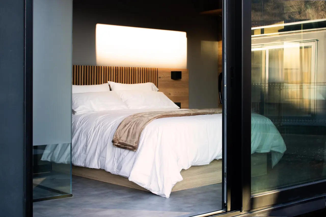 Vista de un dormitorio moderno con cama grande vestida de blanco, cabecero de madera vertical y luz indirecta