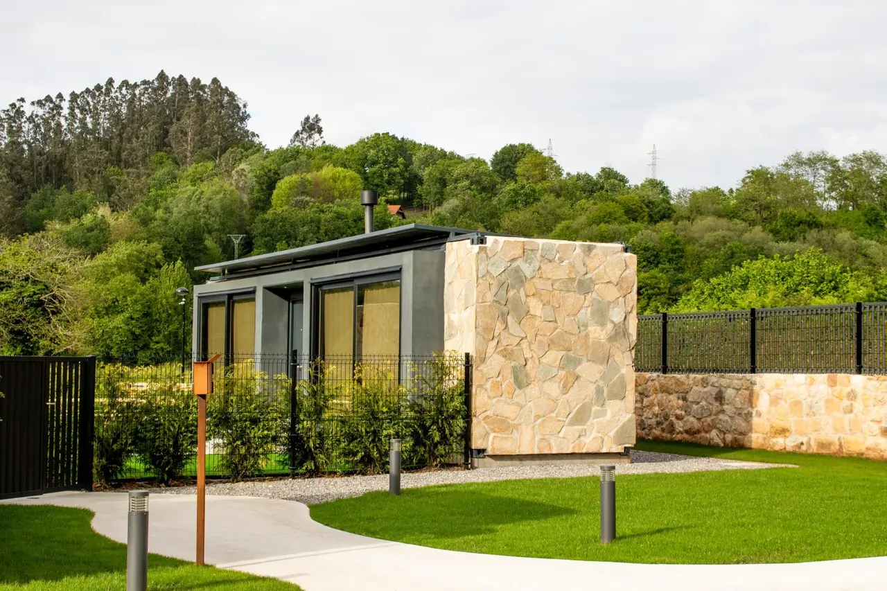 Vista exterior de una pequeña casa moderna con fachada de piedra y paneles oscuros, rodeada de césped, arbustos