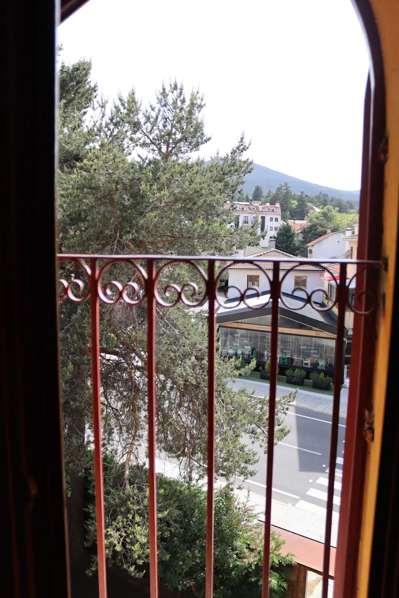 Vista desde una ventana arqueada con barandilla de hierro forjado rojo, mostrando árboles, una calle, edificios
