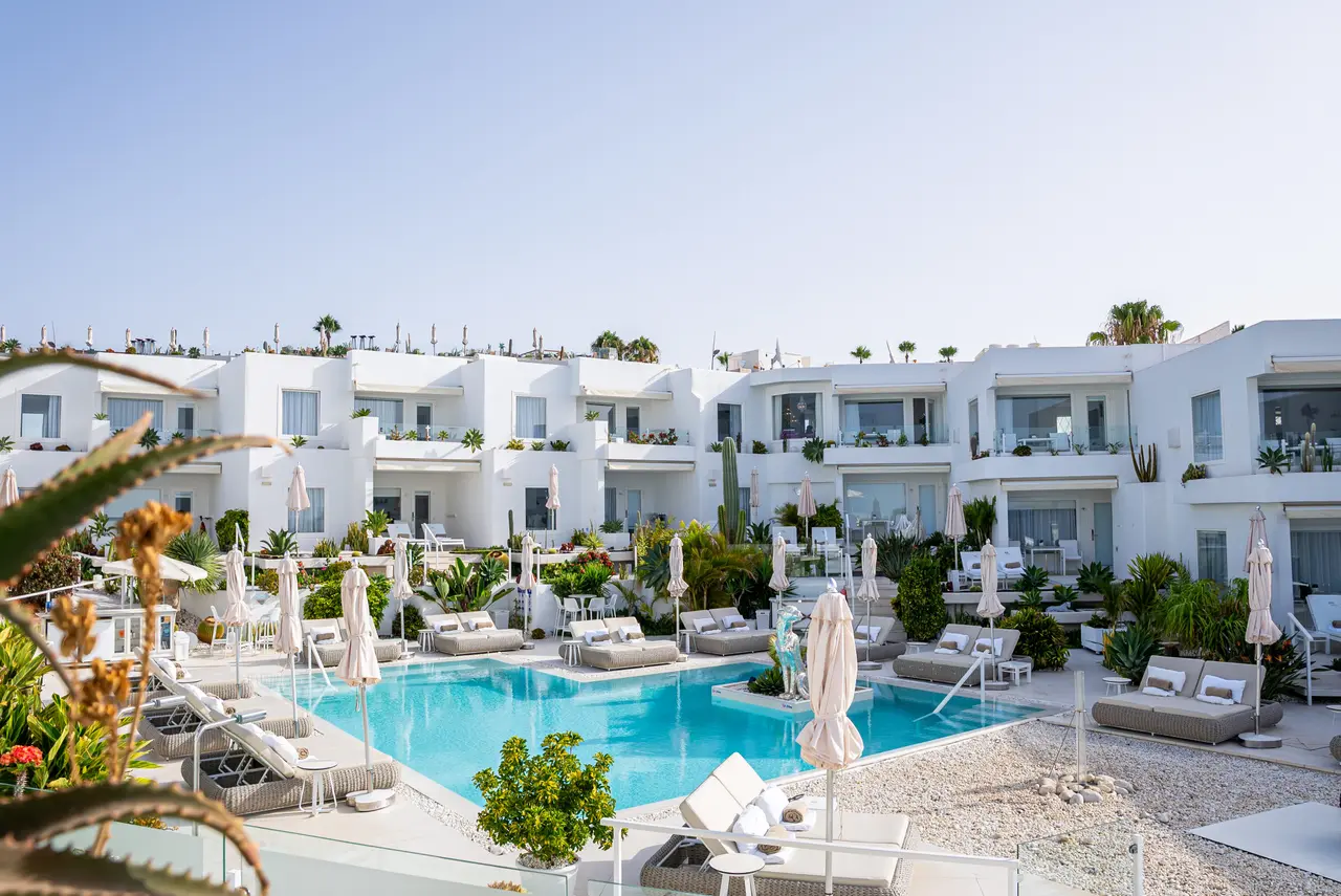 Vista de una piscina rectangular de agua turquesa rodeada de tumbonas, sombrillas cerradas y vegetación, con edificios