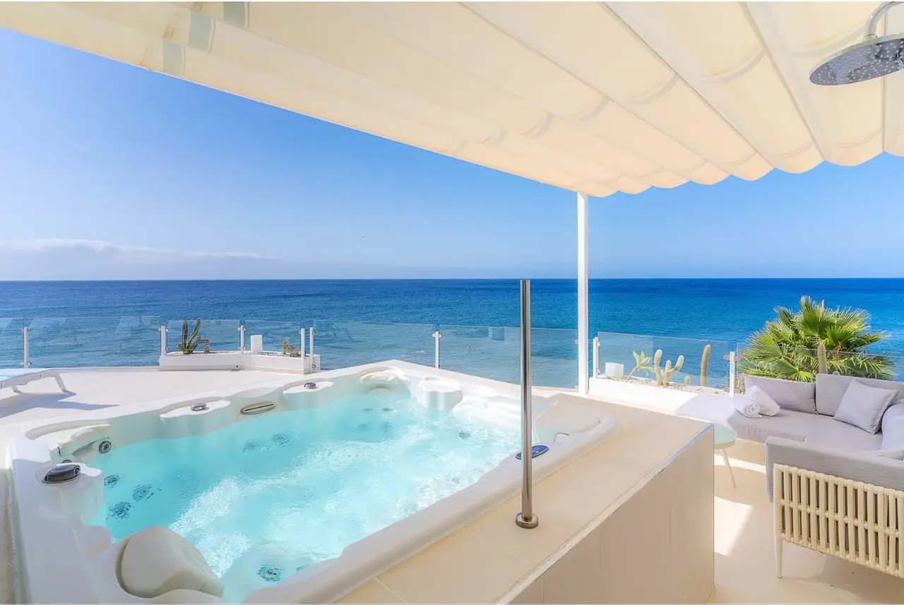 Jacuzzi en terraza privada con vistas al mar, bajo un toldo blanco, con sofá exterior y barandilla de cristal.
