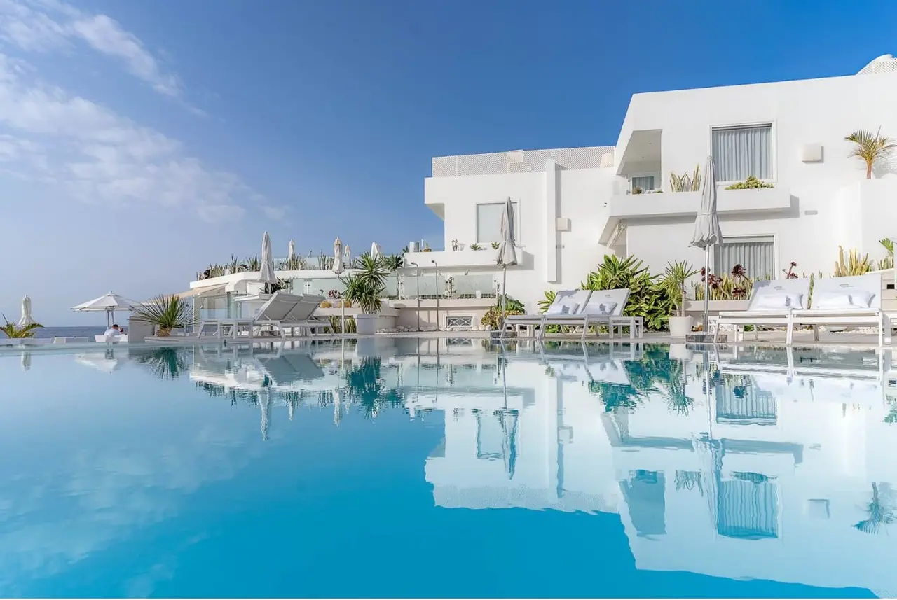 Vista de una piscina infinita con agua turquesa reflejando un edificio blanco moderno de varios niveles, con tumbonas