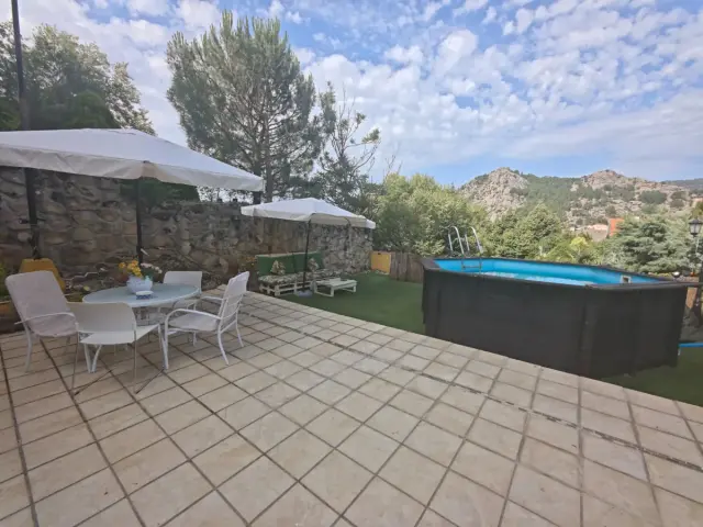 Terraza exterior con suelo de baldosa, mesa y sillas bajo sombrillas blancas, césped, piscina rectangular y bañera