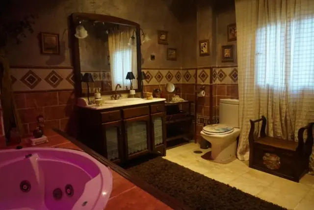 Baño privado con jacuzzi iluminado en color rosa en primer plano, doble lavabo de madera con espejo grande, inodoro