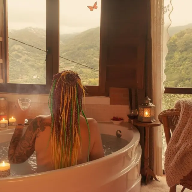 Persona con rastas de colores sentada de espaldas en un jacuzzi redondo dentro de una habitación, con vistas