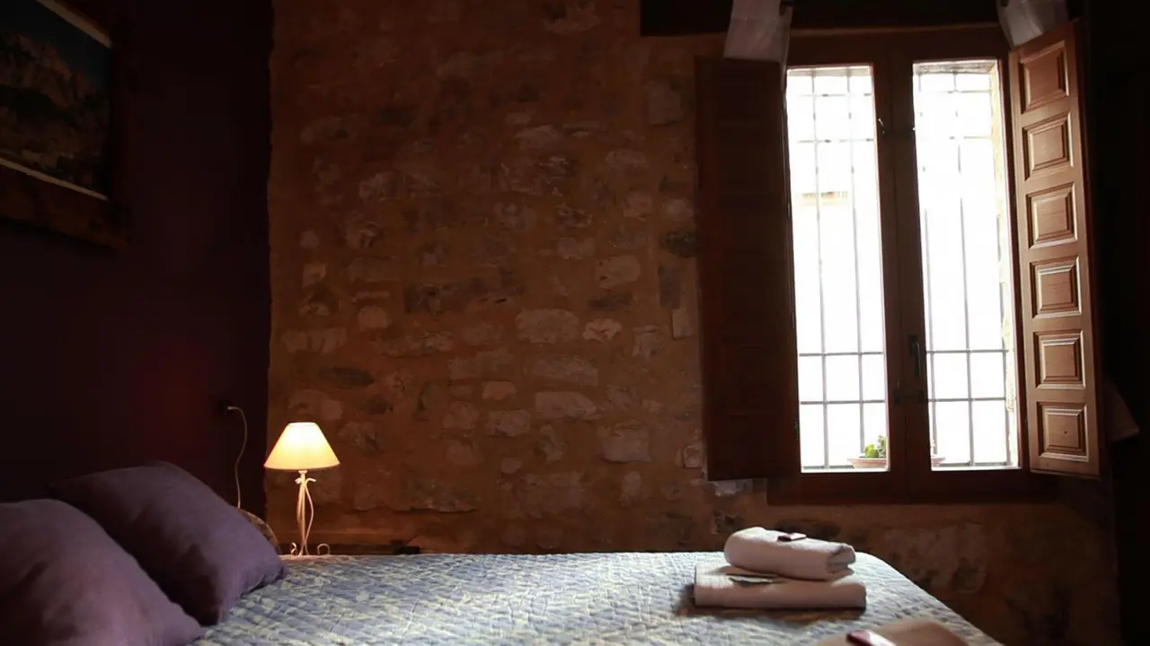 Habitación con cama doble, paredes de piedra vista, ventana de madera abierta y lámpara de pie encendida