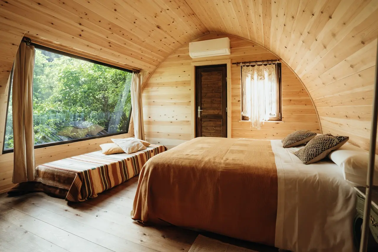Interior de una cabaña de madera con techo abovedado, una cama grande con colcha beige, una cama individual con colcha