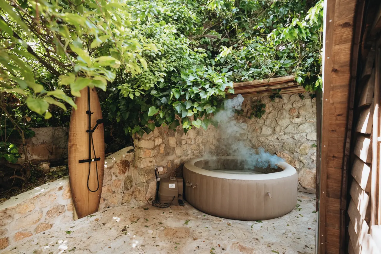 Jacuzzi exterior redondo de color beige, instalado sobre suelo de grava en un jardín privado rodeado de muro de piedra
