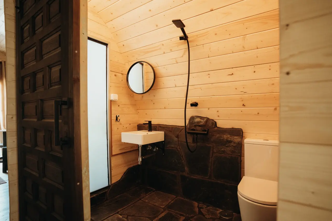 Vista de un baño interior con paredes de madera clara, suelo de piedra irregular, lavabo blanco rectangular