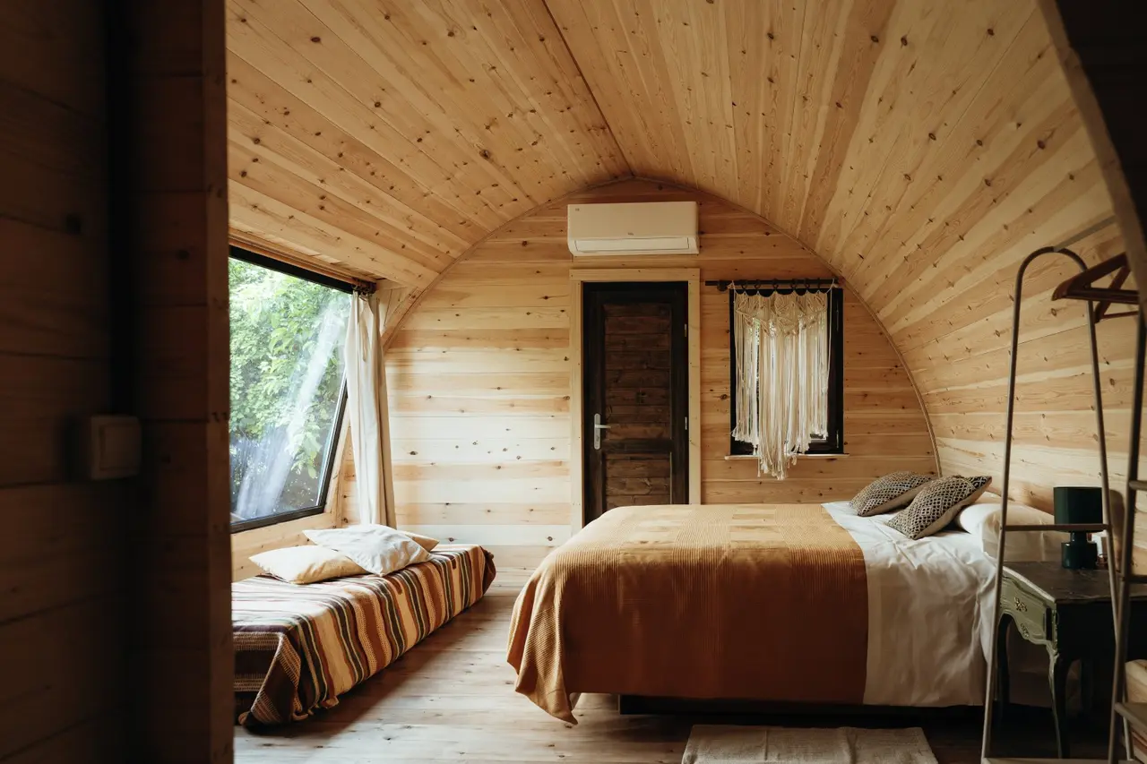 Interior de una cabaña con paredes y techo abovedado de madera clara. En el centro hay una cama doble con colcha