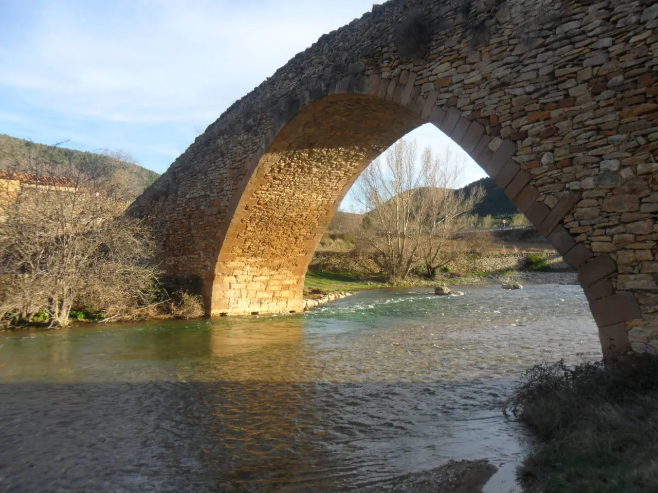Vista de un antiguo puente de piedra sobre un río con agua transparente, rodeado de vegetación y colinas al fondo bajo